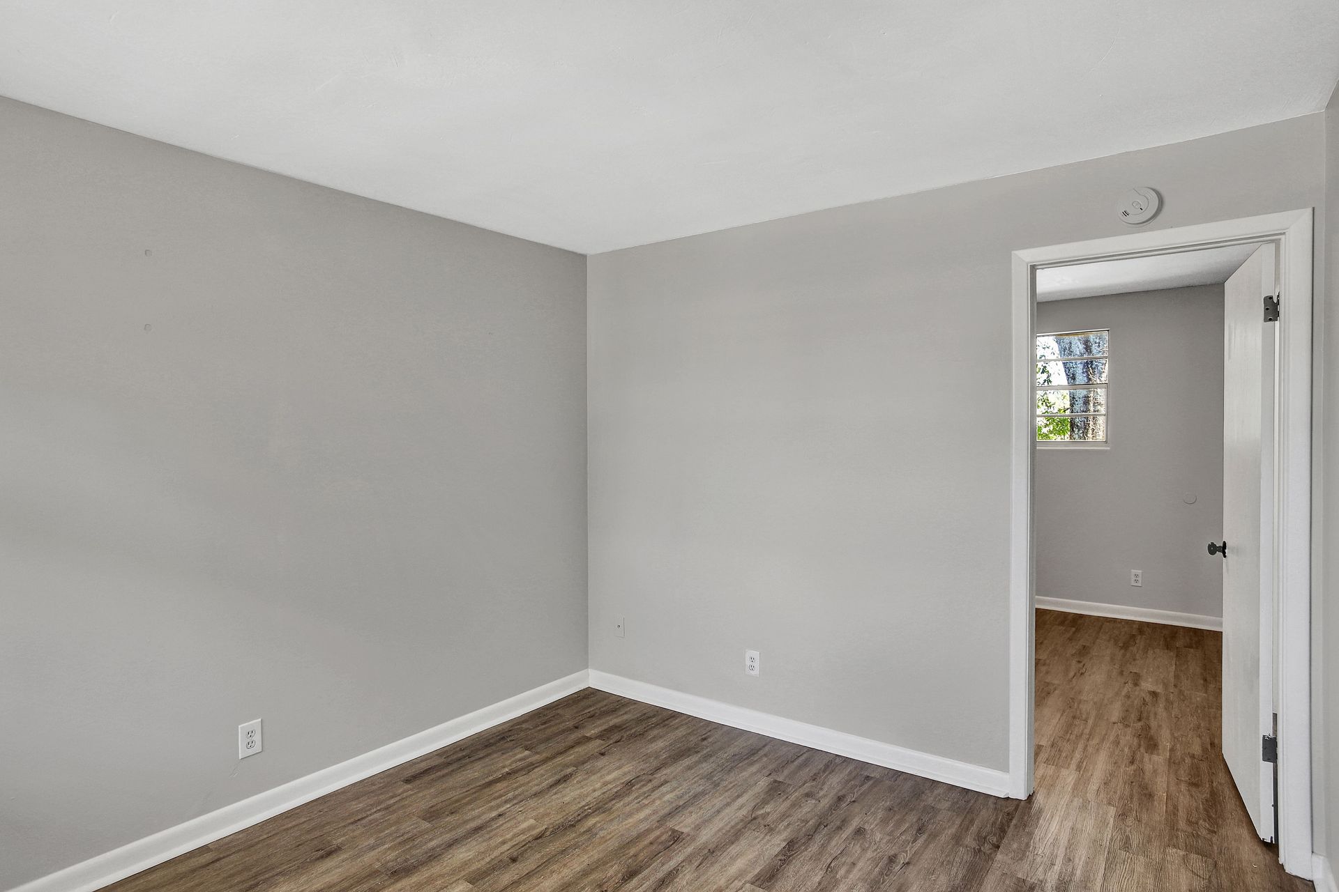 Empty room with gray walls, white trim, and wood-look flooring; doorway to another room with a window.