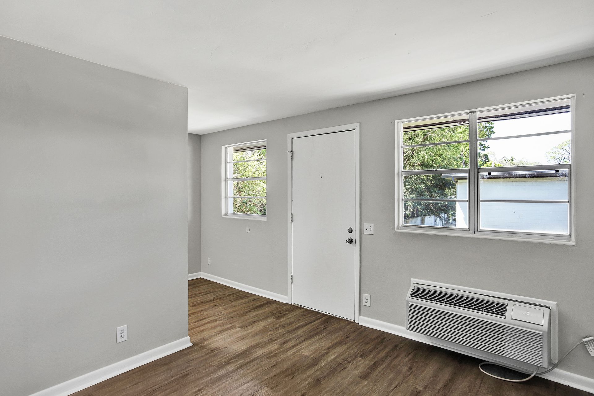 Empty room with gray walls, wood-look flooring, door, two windows, and an air conditioner.
