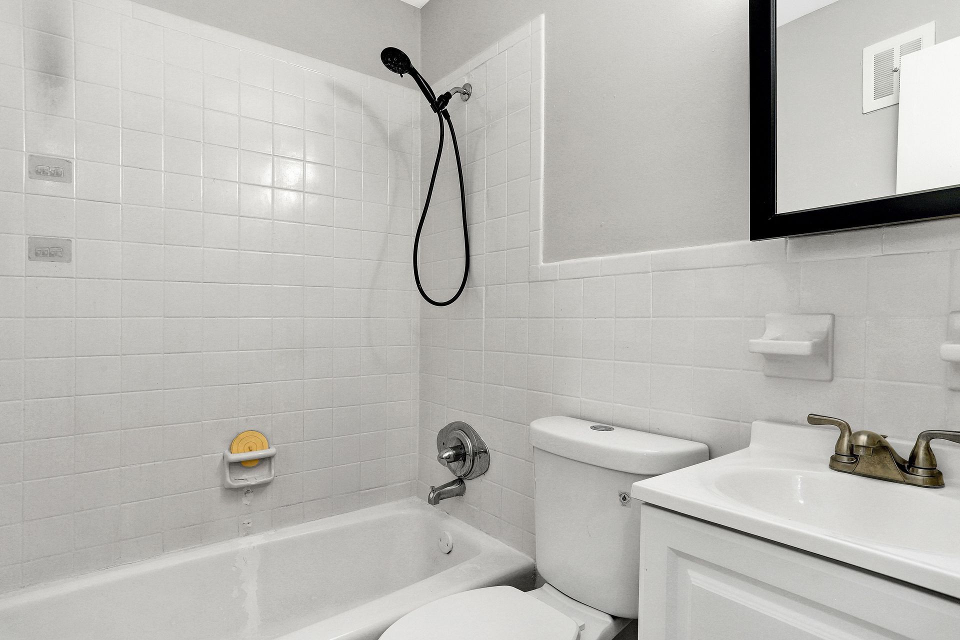 White tiled bathroom with a tub, toilet, and sink. Black showerhead and mirror.