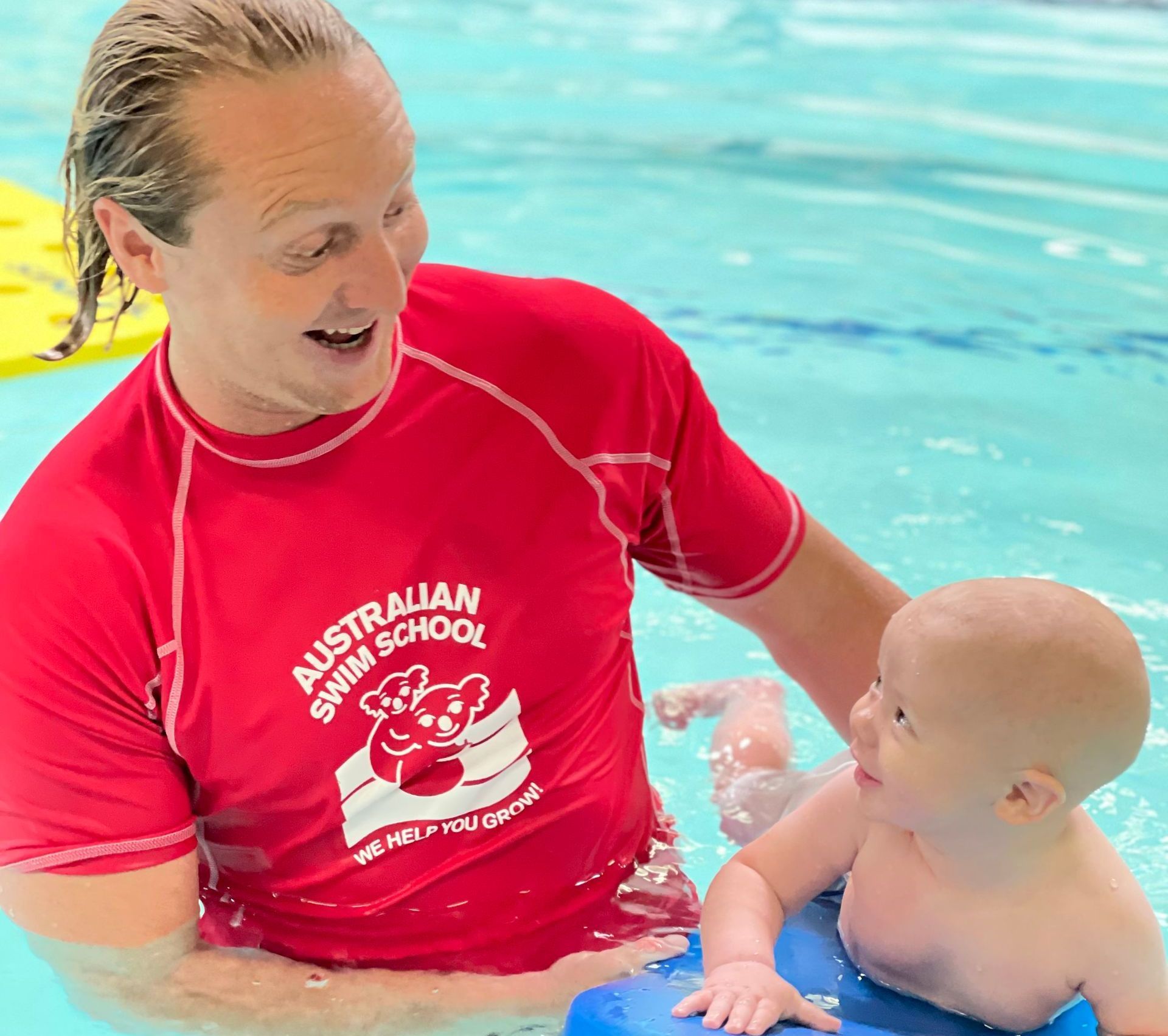 A man in a red australian swim school shirt is playing with a baby in a pool