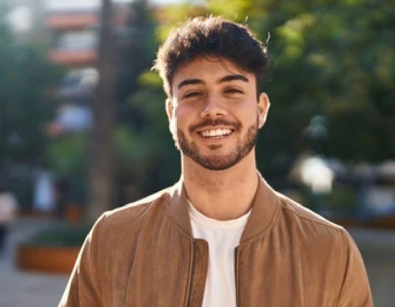 A young man with a beard is smiling for the camera while wearing a brown jacket.