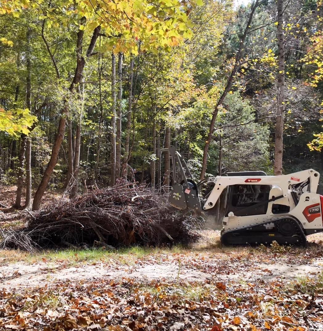 A white skid steer with tracks clearing a large pile of brush in a wooded area during autumn.