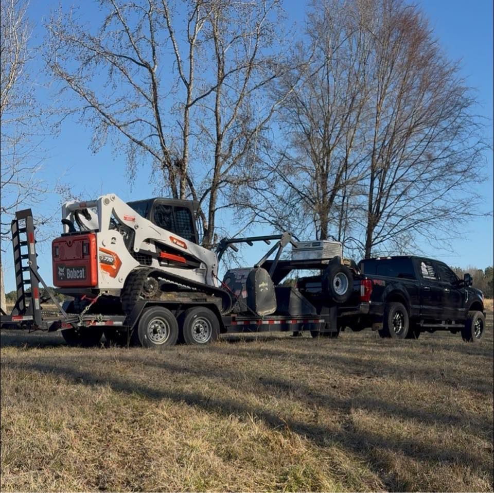 A black pickup truck parked in a field, towing a flatbed trailer loaded with a white and orange Bobcat skid-steer loader.