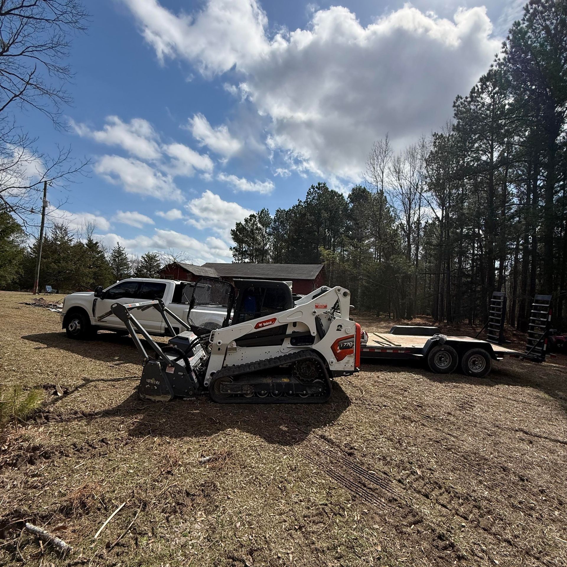 A white skid steer with a forestry mulcher attachment sits in a field near a white pickup truck and a flatbed trailer.