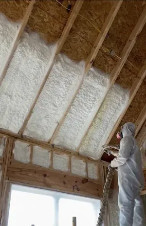 A person in a protective suit and respirator sprays white insulation foam onto wooden rafters in a building under construction.