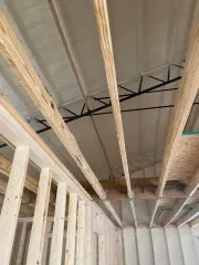 Framing of a room under construction, showing wooden wall studs, floor joists, and exposed steel roof trusses.