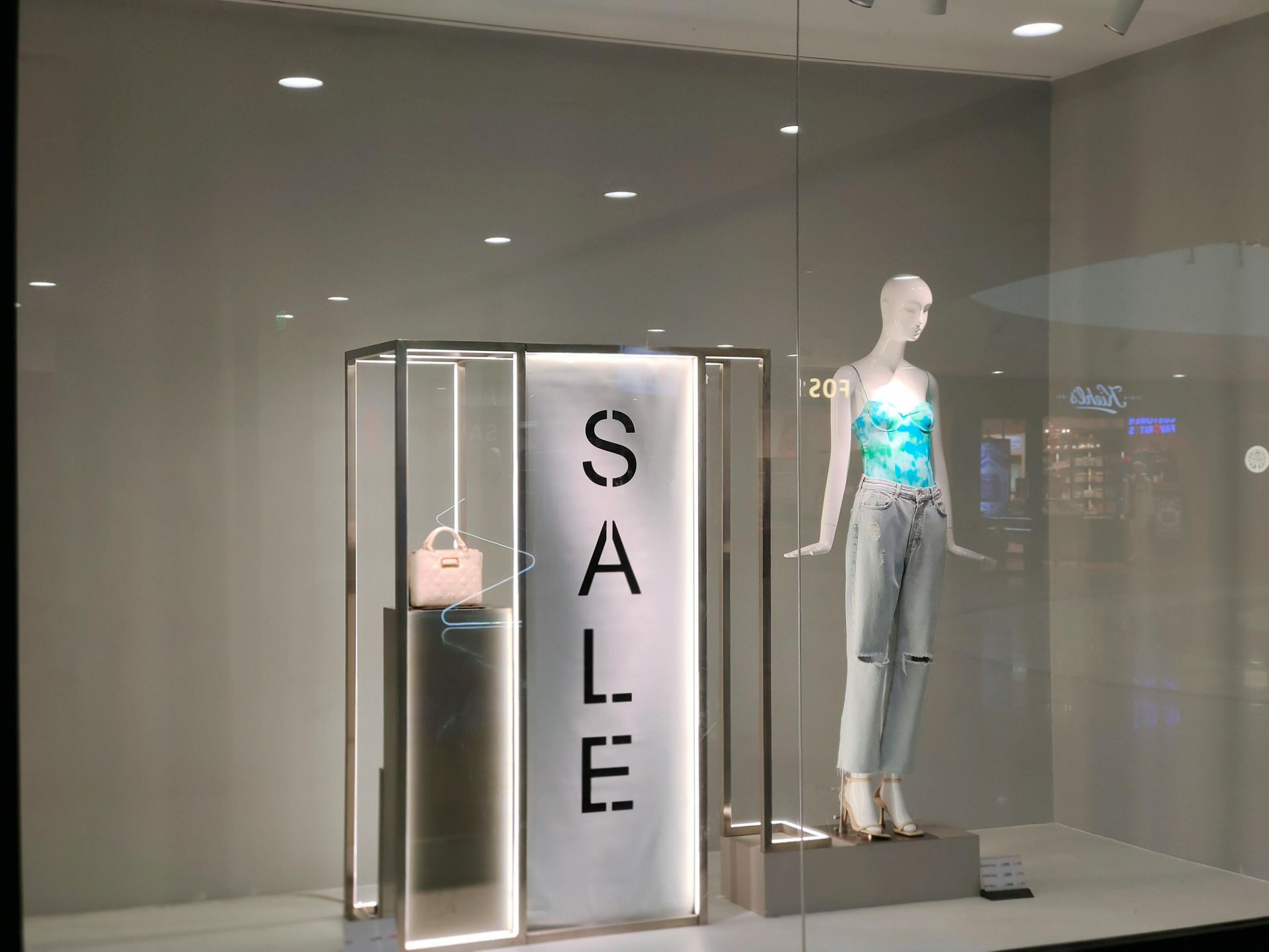 Store window display with a SALE sign, handbag, and mannequin in a blue top and jeans.
