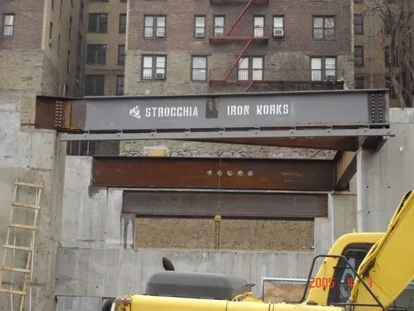 A Yellow Excavator Sits Under A Bridge That Says Iron Works — Albertson, NY — Patsy Strocchia & Sons Ir

