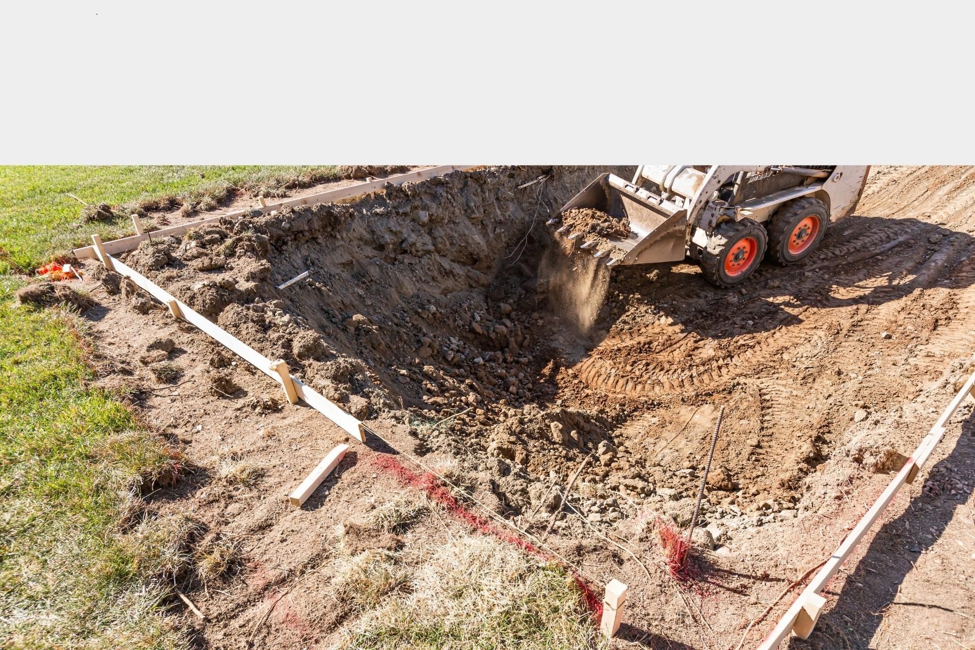A skid steer loader excavates dirt within a marked construction area with wooden framing boards on a grassy site.