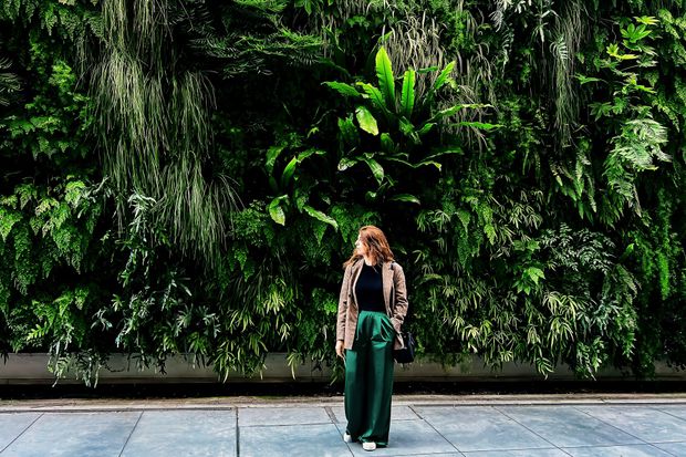 Woman in green pants and patterned jacket stands in front of a lush green wall of plants.