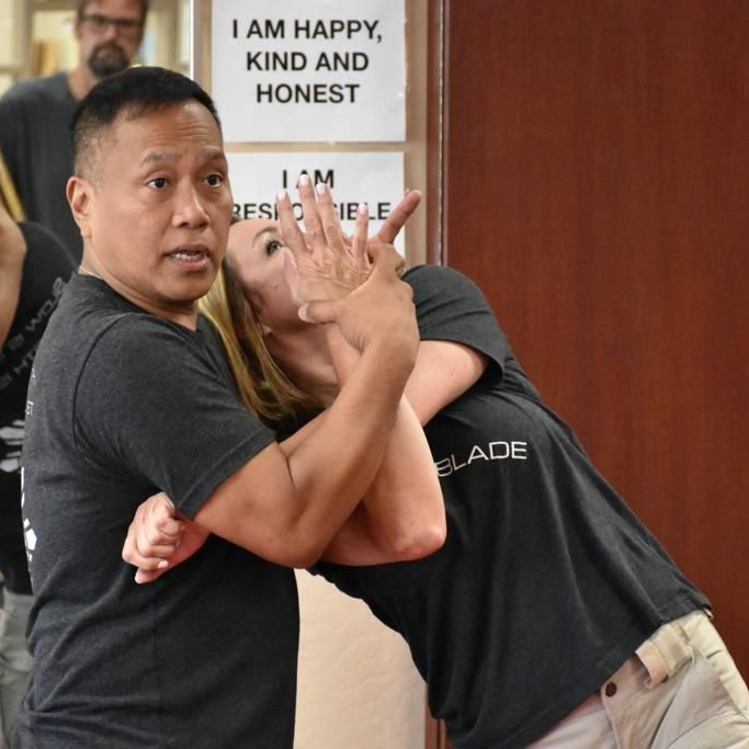 Two men in grappling position on a mat. One embraces the other from behind, both smiling. Gym setting.