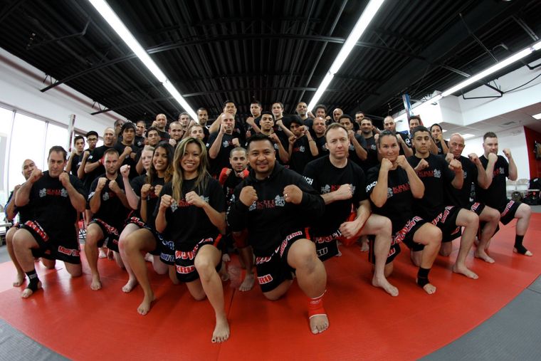 Family in Jiu-Jitsu gis poses. Indoors, blue mats, a smiling woman in the middle, around a seated man with a beard.