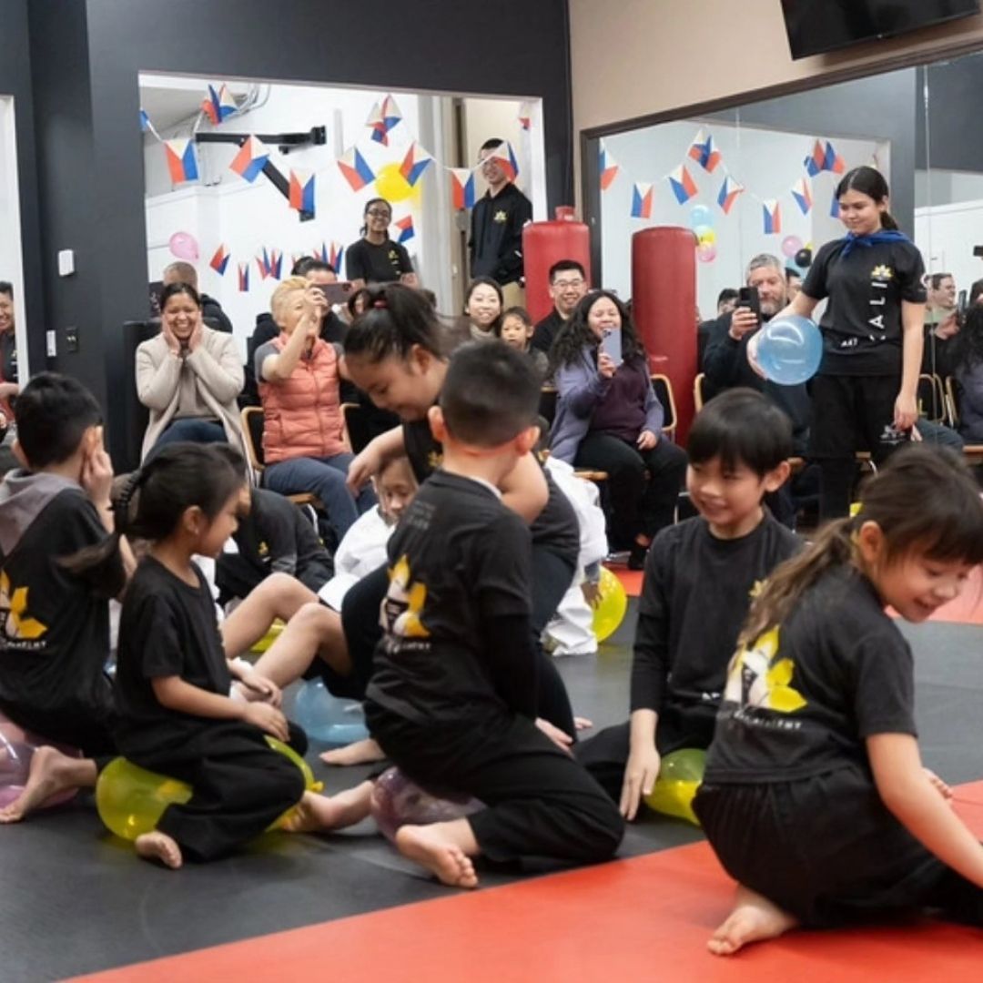 Two children practicing Brazilian Jiu-Jitsu on a mat. One child in white gi has the other in a leg lock. A girl watches.