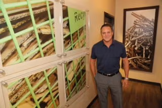 Man standing beside a large framed close-up artwork in a hallway with wood flooring and framed photos.