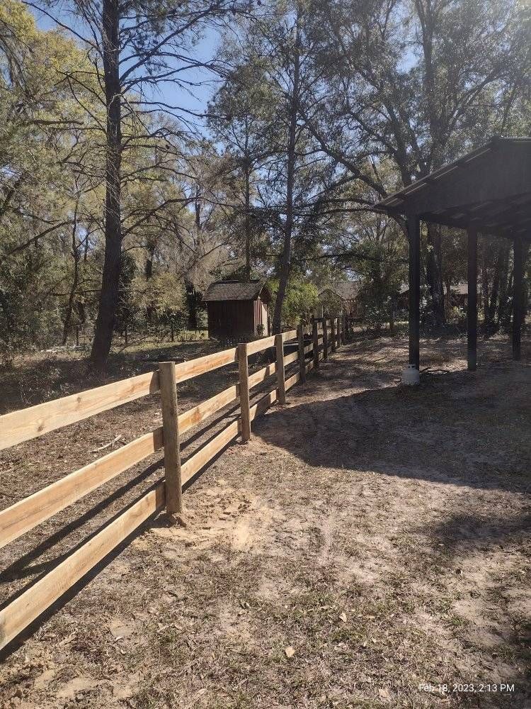 Wooden fence bordering a sandy path; trees and a pavilion in the background. Sunny day.