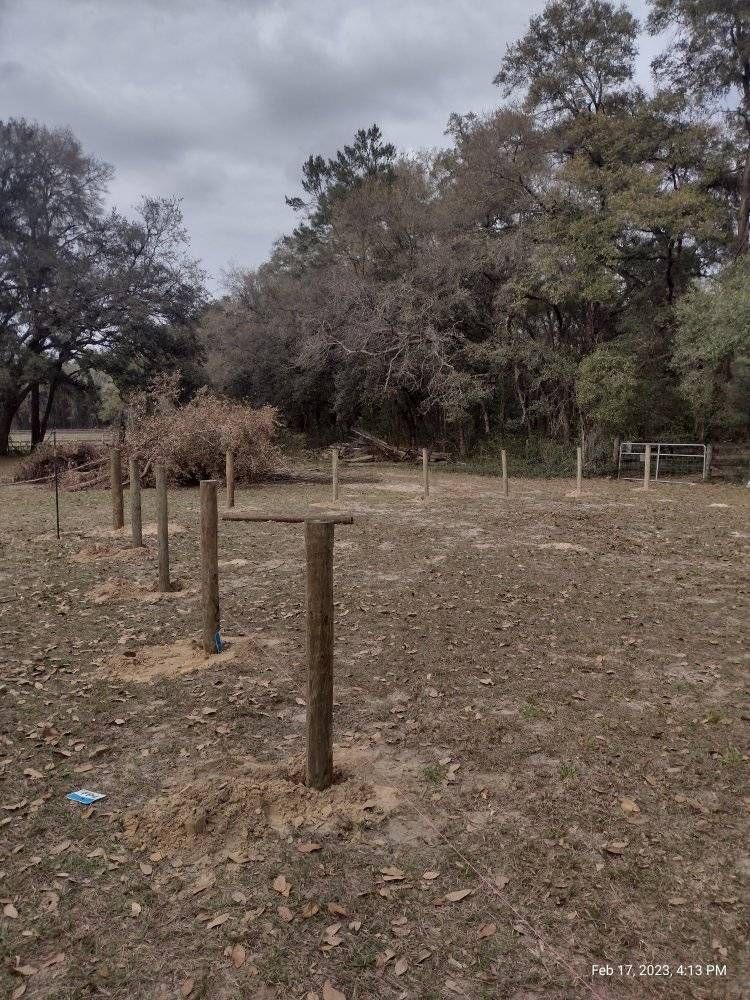 Wooden fence posts set in a grassy field, trees in the background under a cloudy sky.