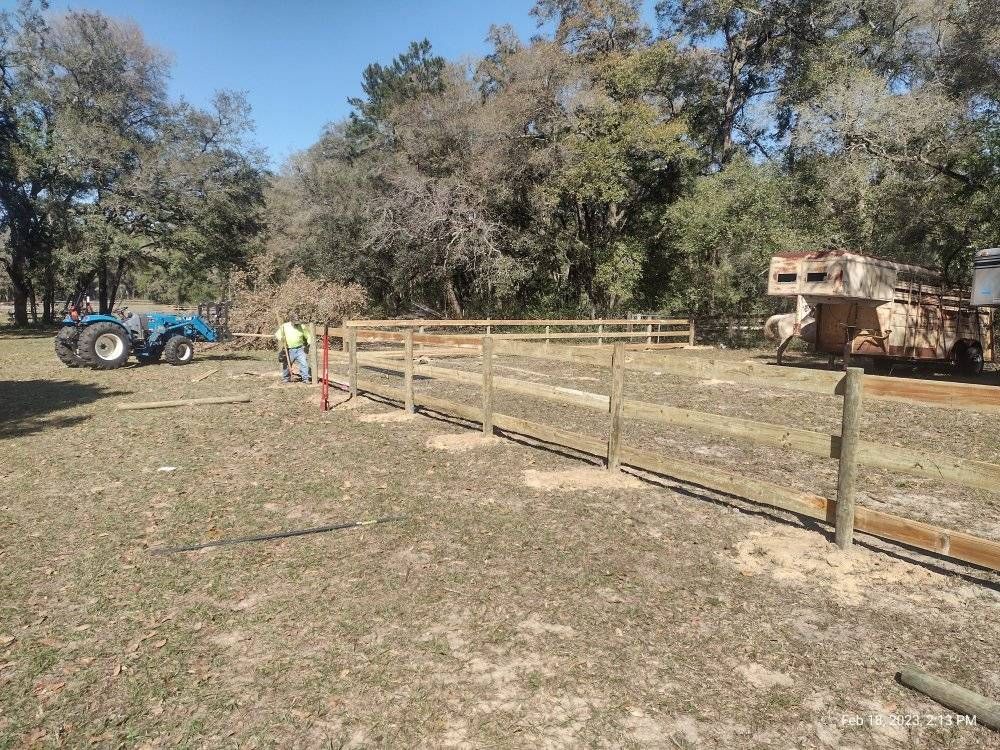Fence being built in a grassy field. Two people working, tractor, trailer, and trees in the background.