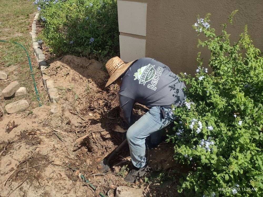 Man in hat digging in dirt near a building, next to a bush.