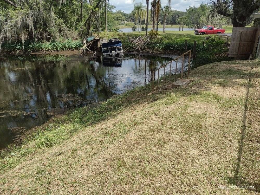 Truck submerged in pond, grass embankment in foreground, trees and red truck in background.