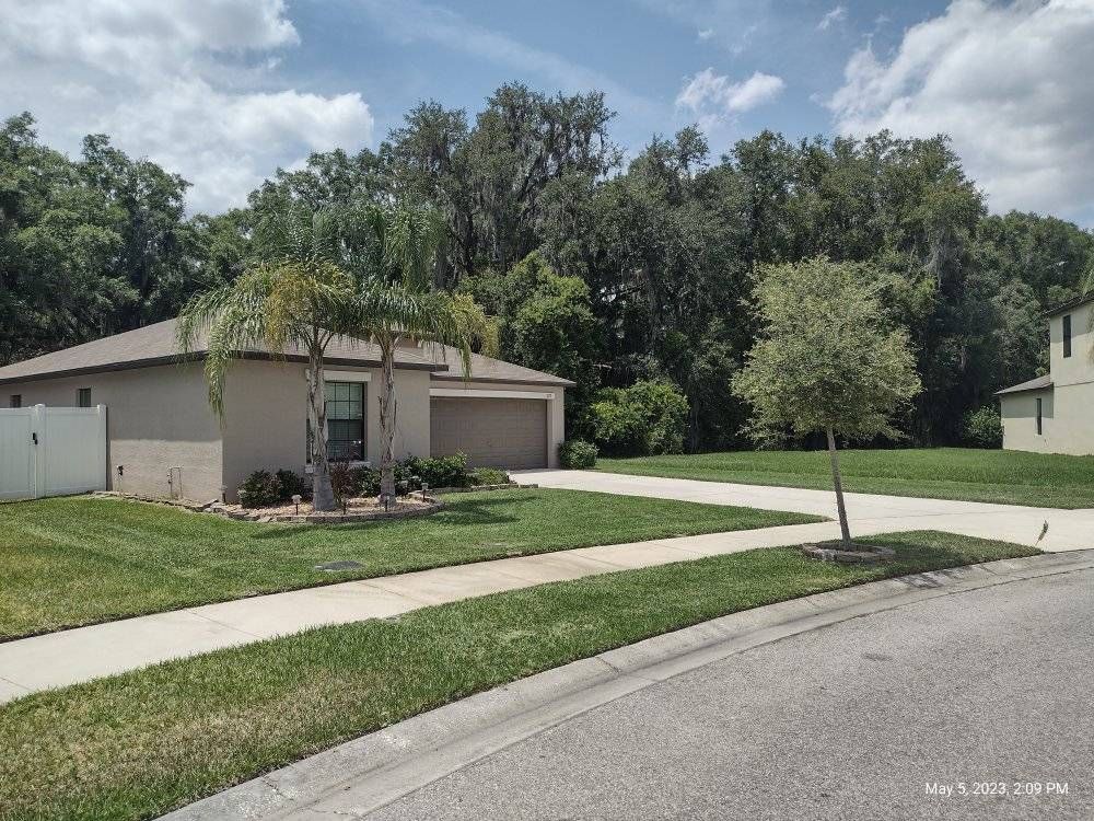 Tan house with attached garage, green lawn, sidewalk, and trees in background.