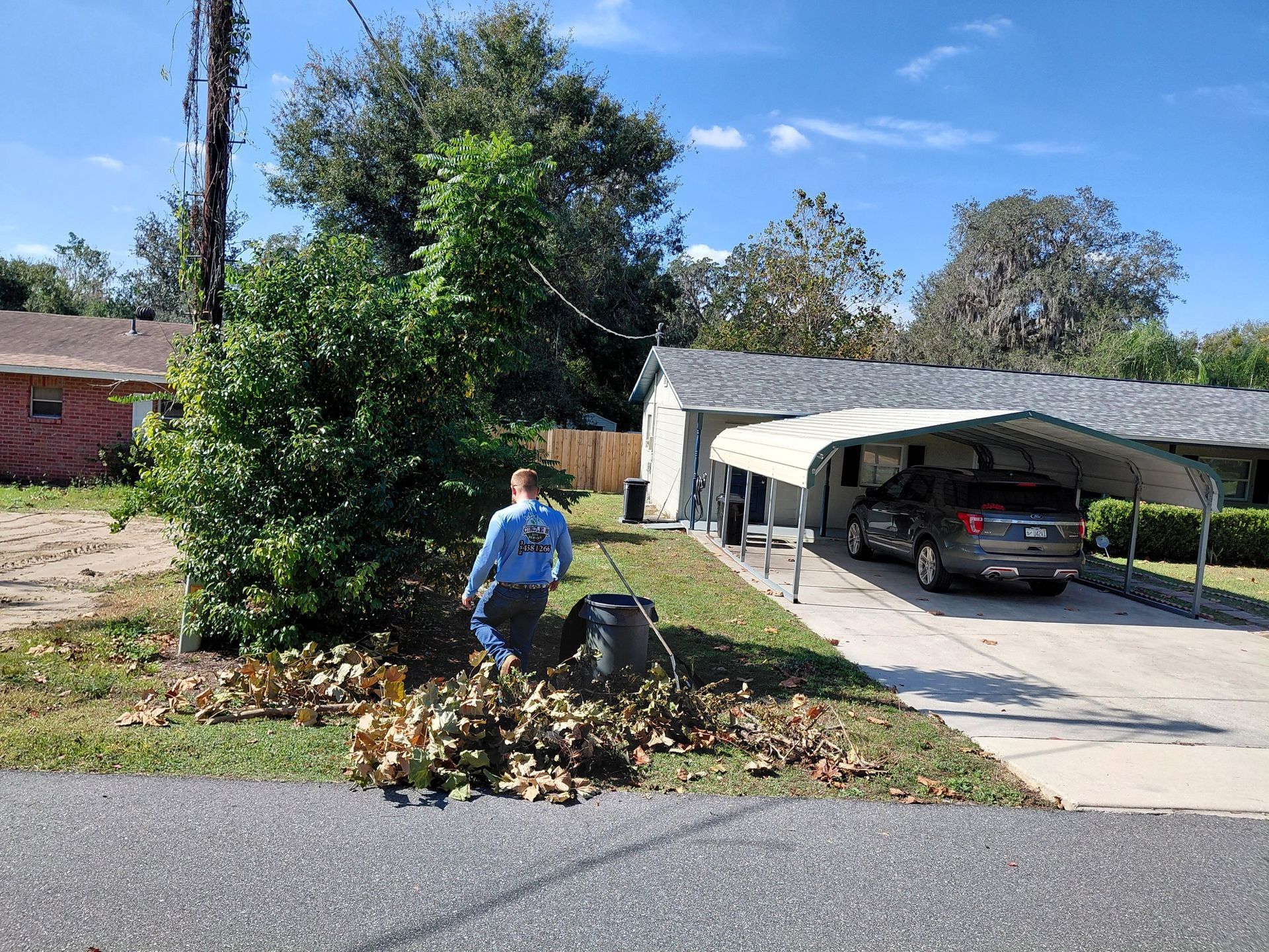 Person raking leaves in front yard; house with carport in background. Blue sky and sun.