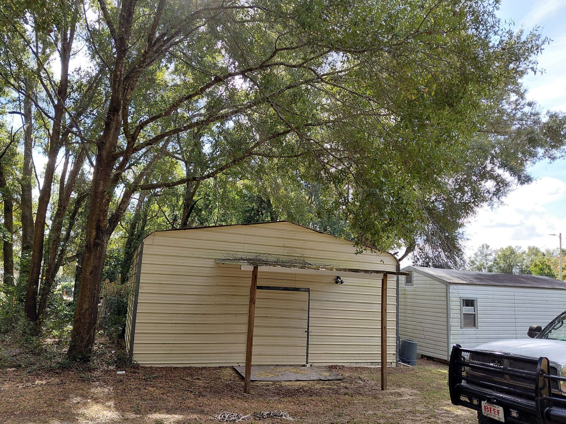 Metal shed with overhang, set in a yard, under a tree. Another shed and truck are visible.