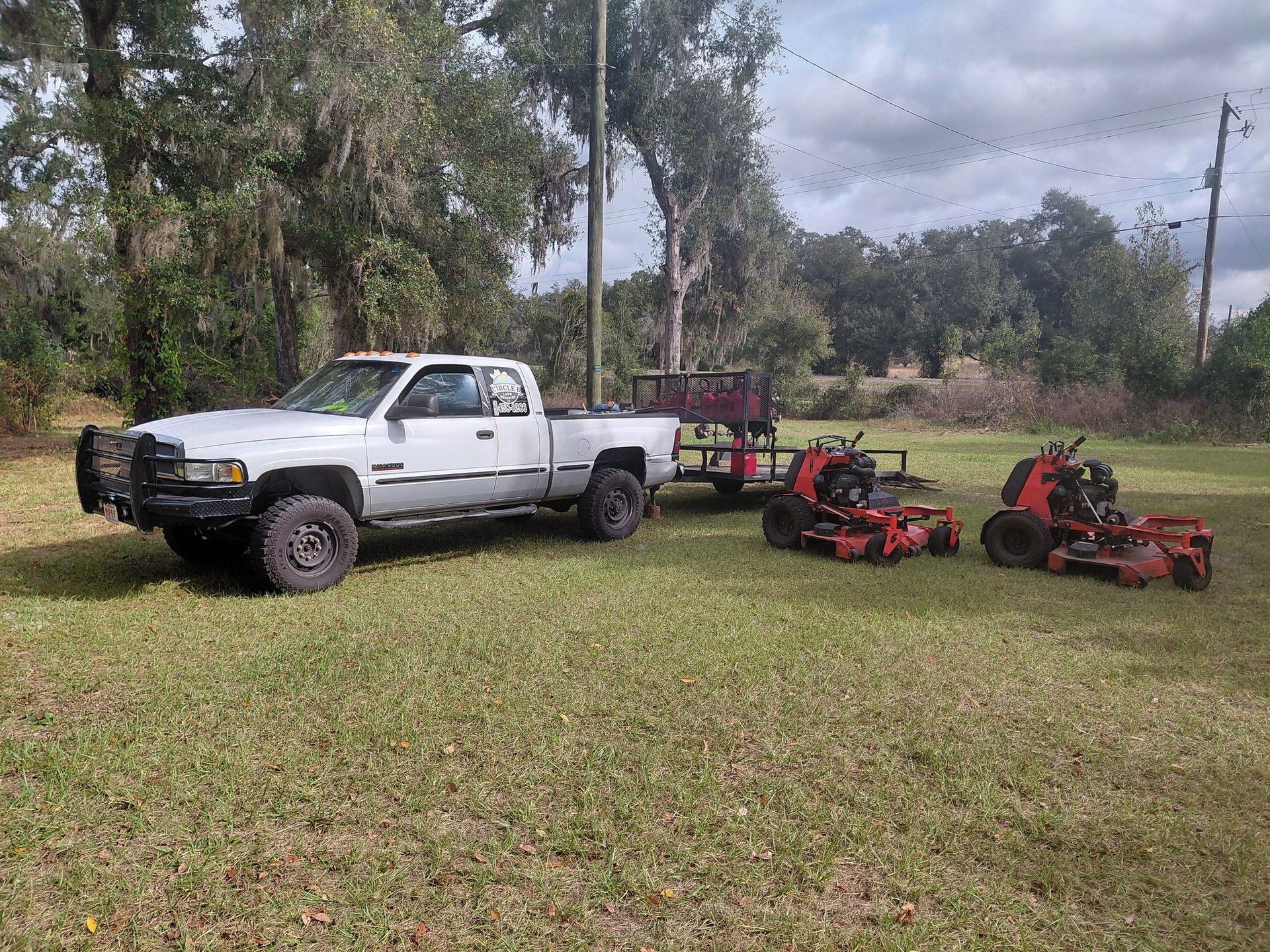White pickup truck towing a trailer with two red lawn mowers on a grassy field.