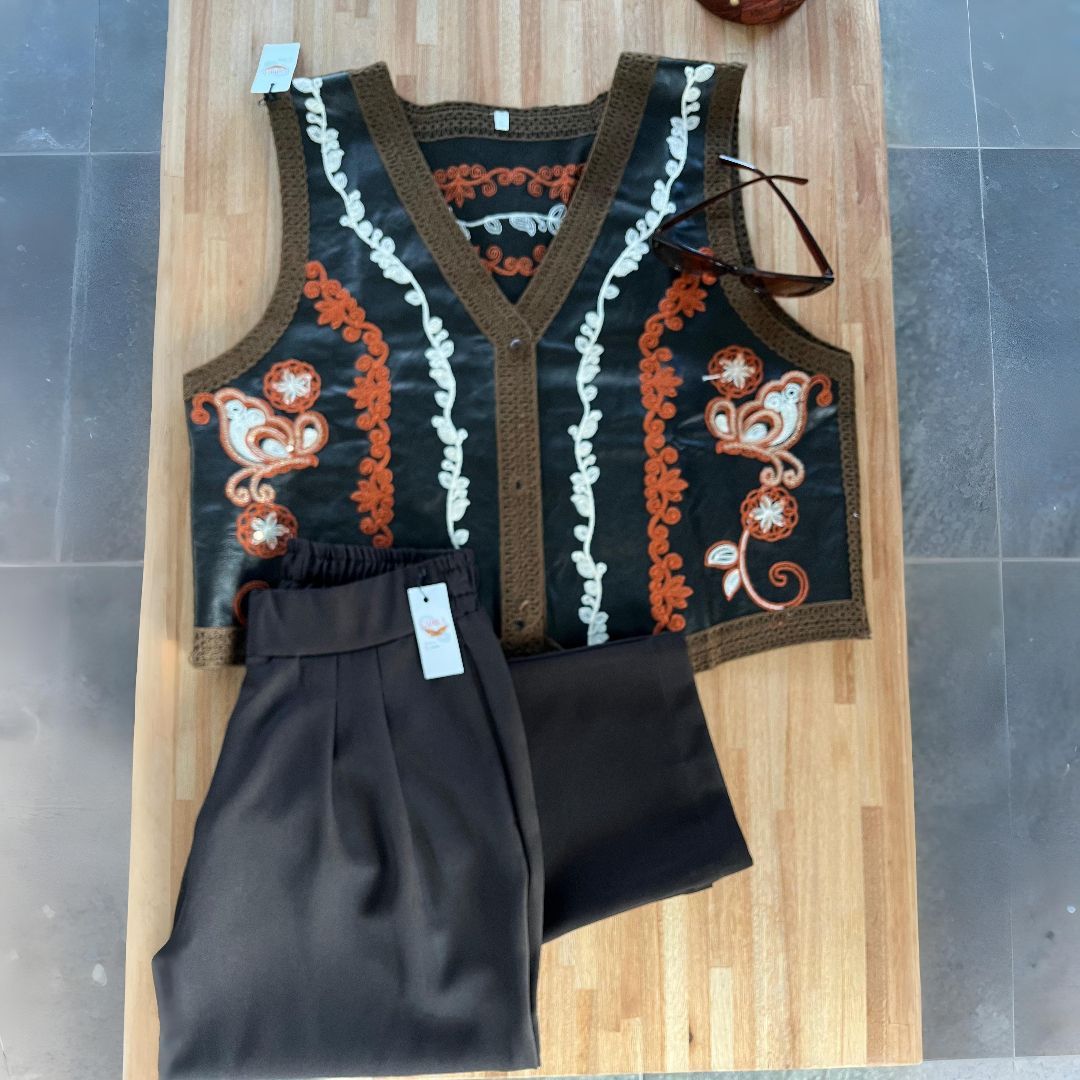 Pantalón color canela y chaleco bordado color crema con ribete marrón sobre superficie de madera.