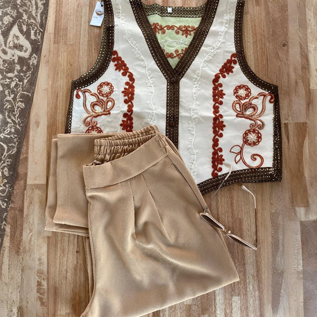 Pantalón color canela y chaleco bordado color crema con ribete marrón sobre superficie de madera.