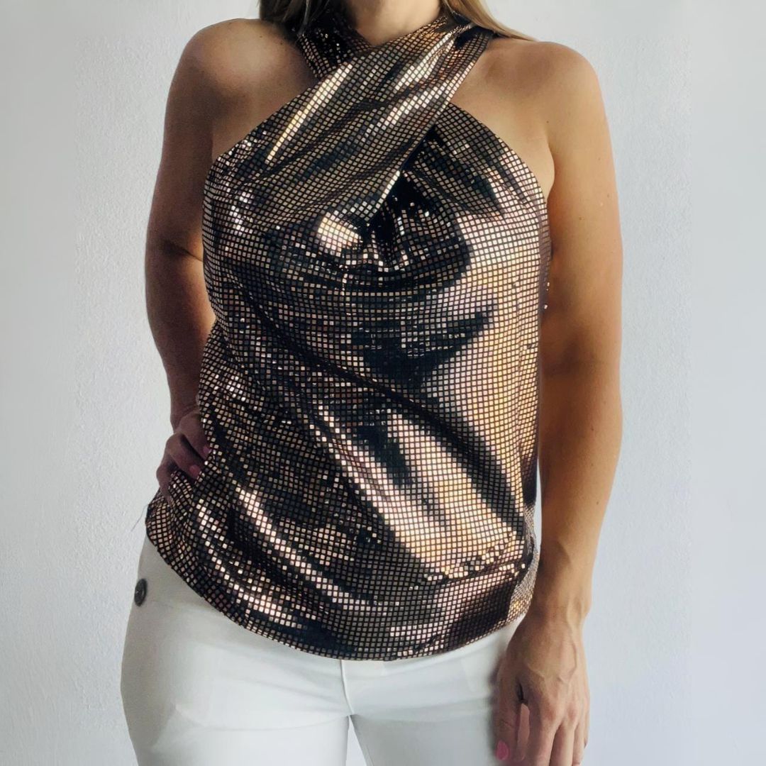Mujer con top dorado cruzado y pantalones blancos. Está de pie con la mano en la cadera.