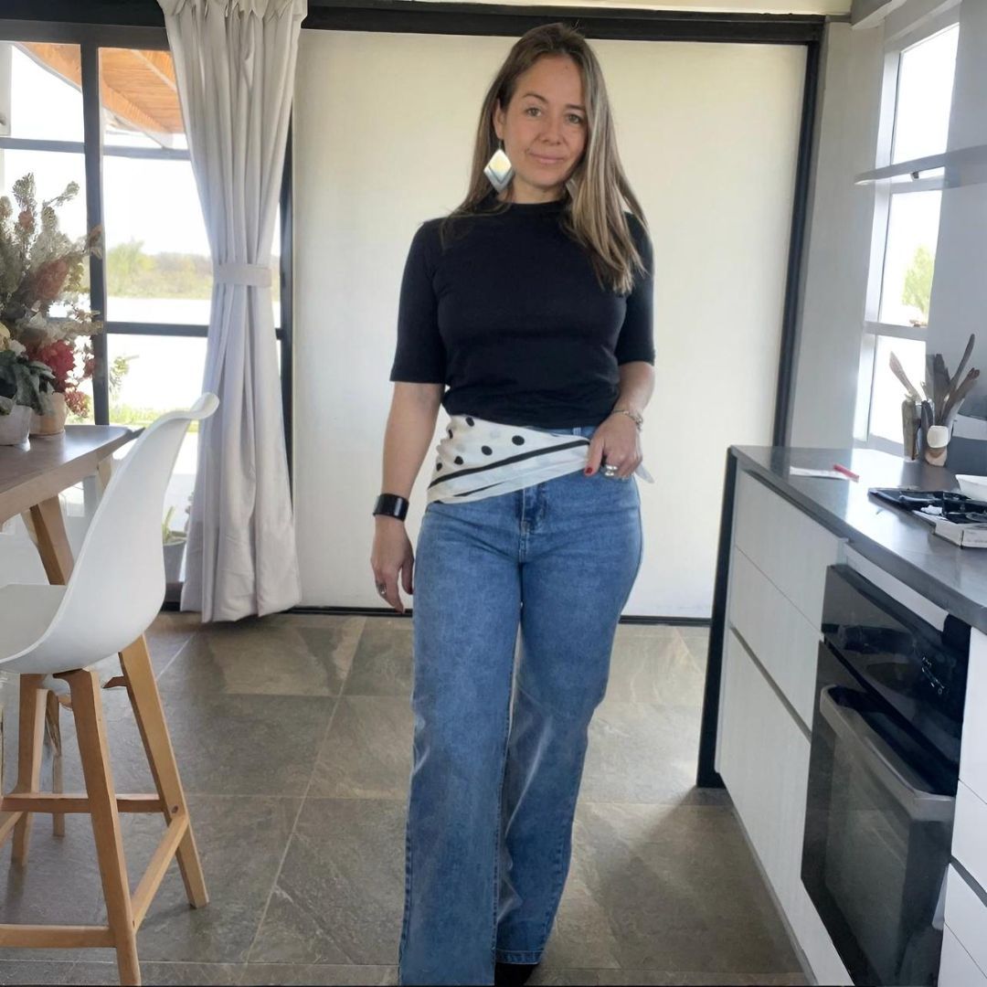 Mujer con top negro y jeans azules, manos en los bolsillos, en el interior.