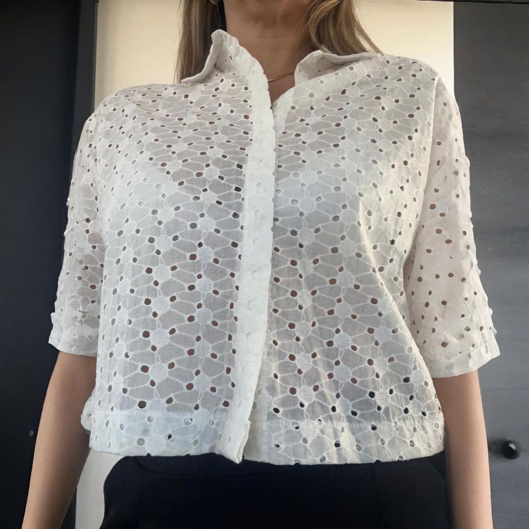 Mujer con top blanco de ojales y mangas cortas, pantalones negros. Interior, fondo neutro.