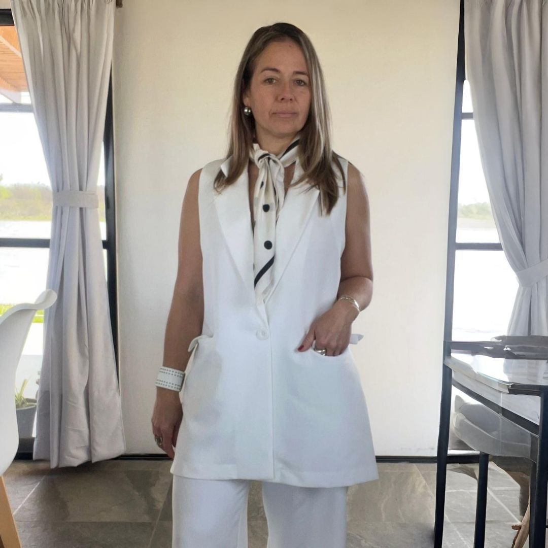 Mujer con chaleco y pantalones blancos, bufanda estampada, de pie en el interior.