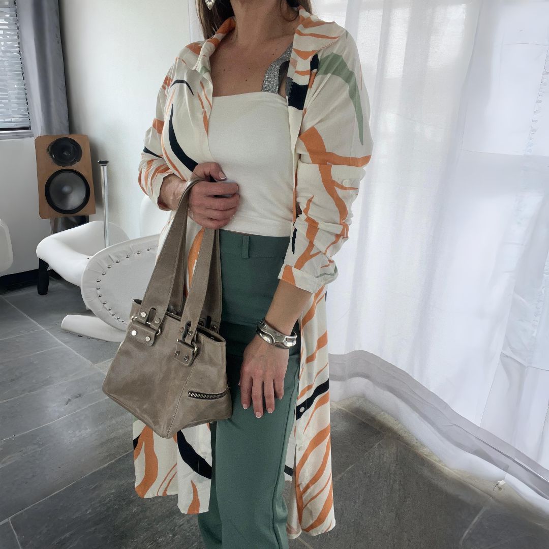 Mujer con camisa larga estampada, top blanco, pantalones verdes, llevando un bolso color canela, de pie en el interior.