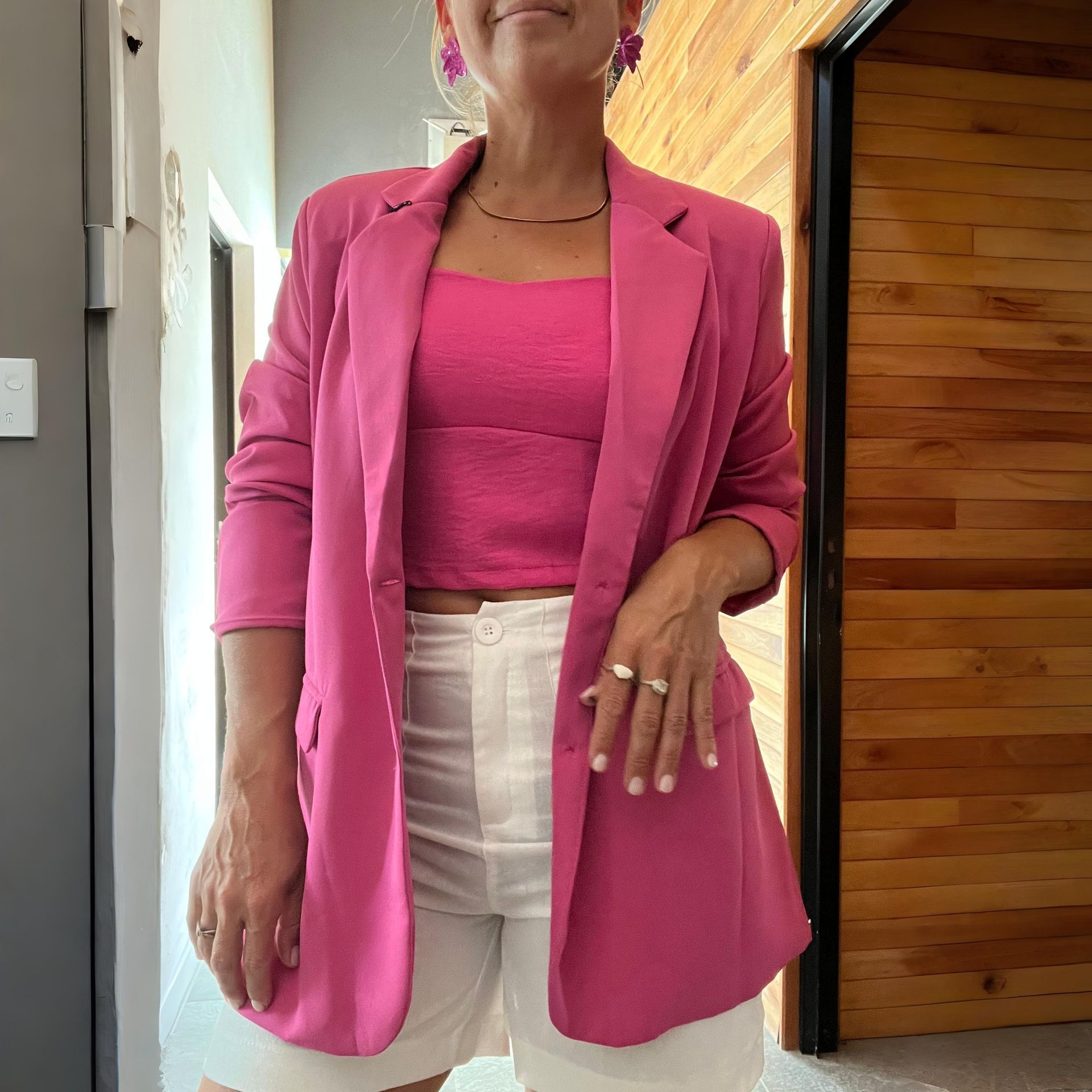 Mujer con blazer rosa, top y pantalones cortos blancos, de pie frente a una puerta de madera.
