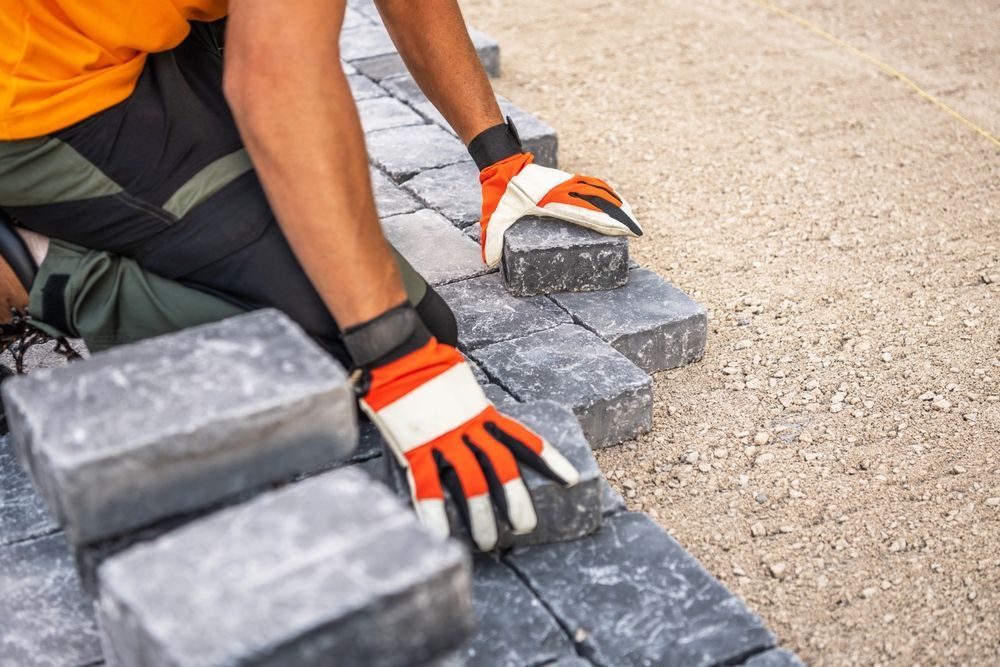 Person wearing orange gloves, laying gray paving stones on a gravel surface.