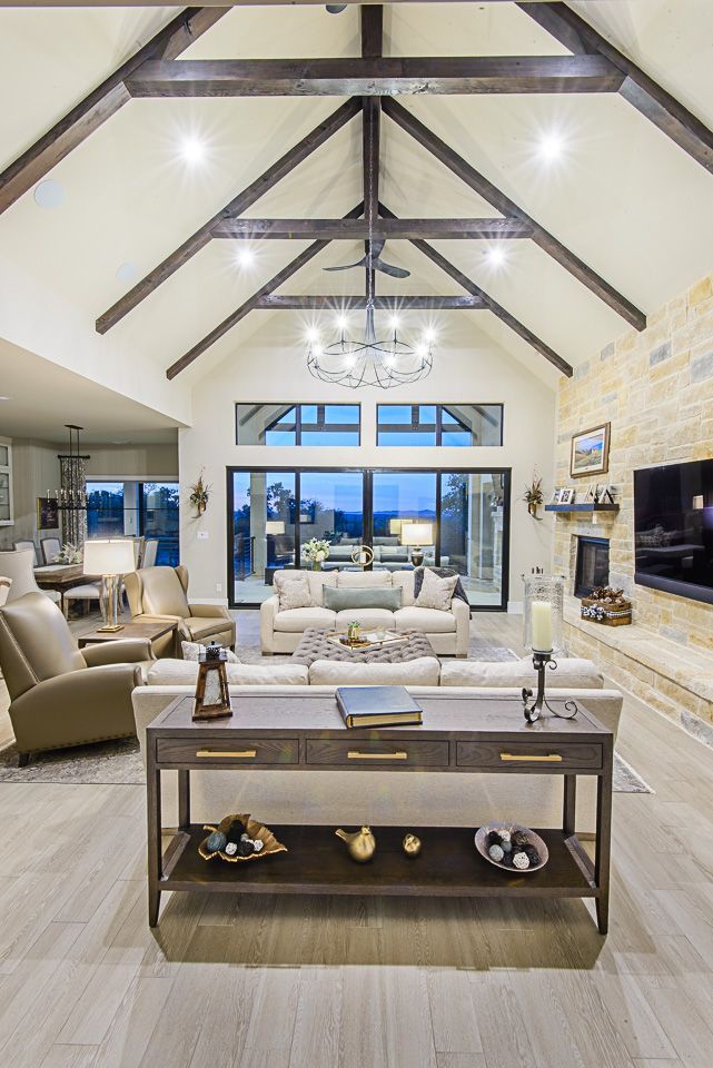 Bright living room with vaulted wood beams, chandelier, large windows, fireplace, and neutral furniture