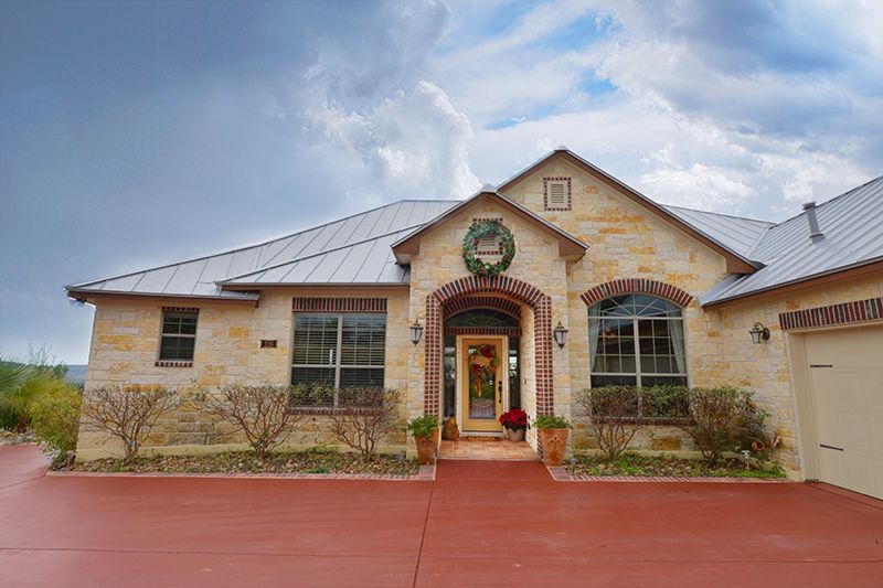 Beige stone house with red driveway under cloudy sky.