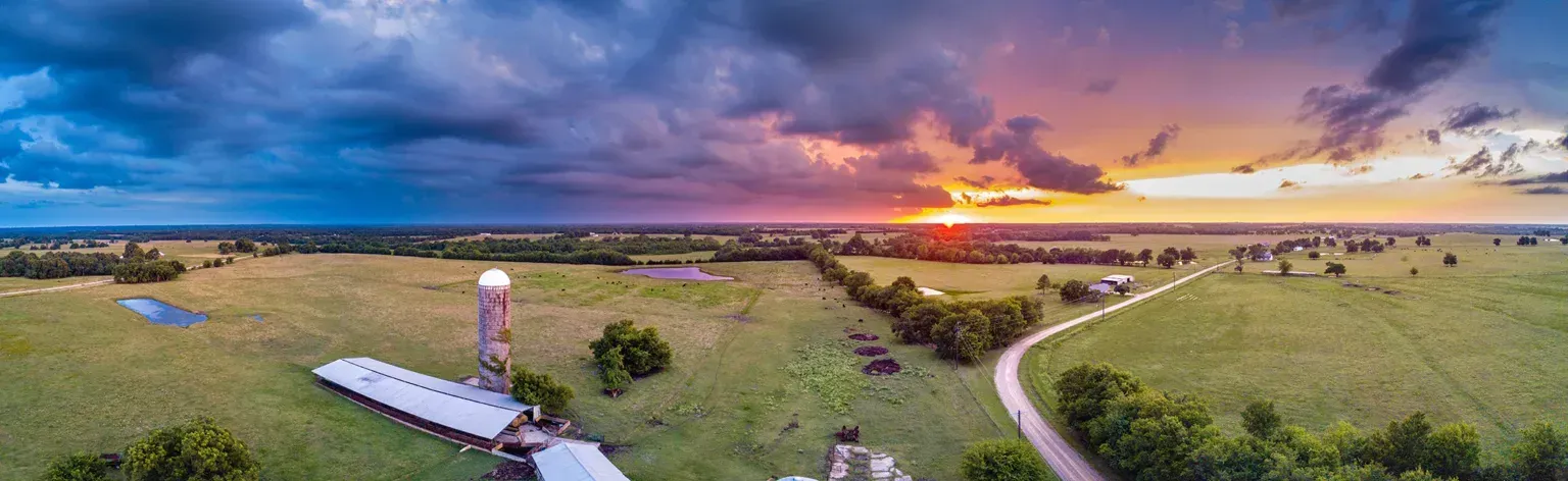 Panoramic aerial view of a farm under a dramatic sunset sky. Fields, buildings, and a long driveway are visible.