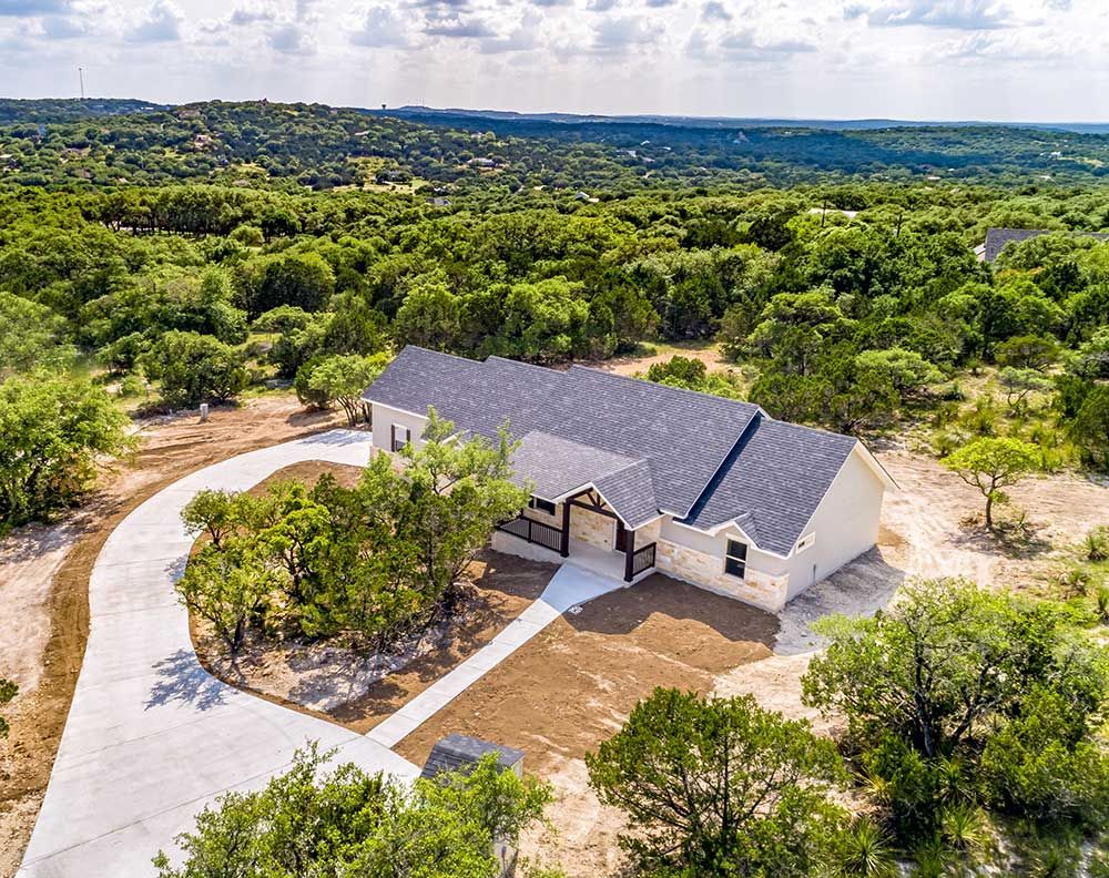 Aerial view of a single-story house with a long driveway surrounded by green trees and a distant landscape.