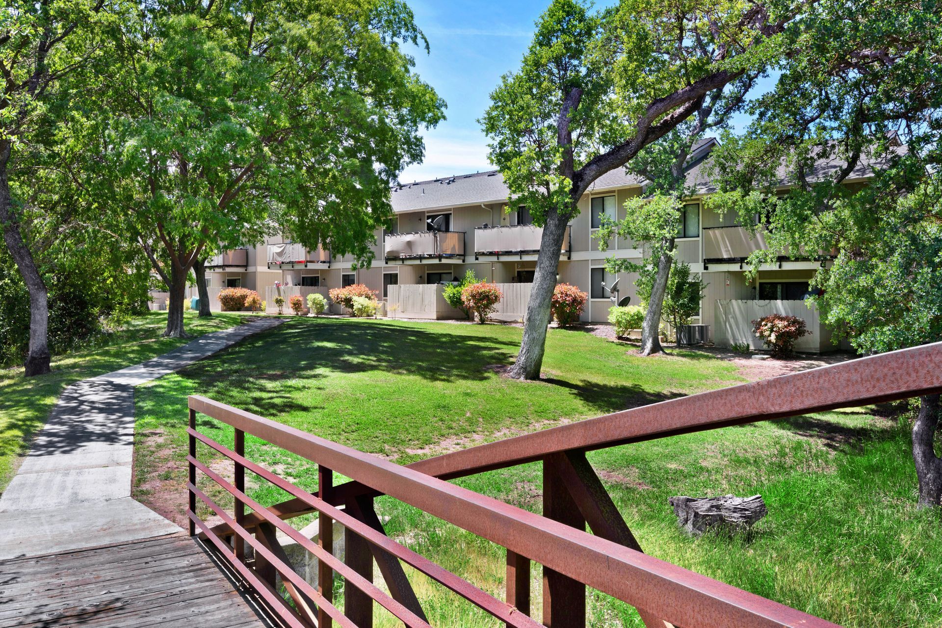 Brown bridge over a grassy area with apartments in the background, trees and a walking path on the left.