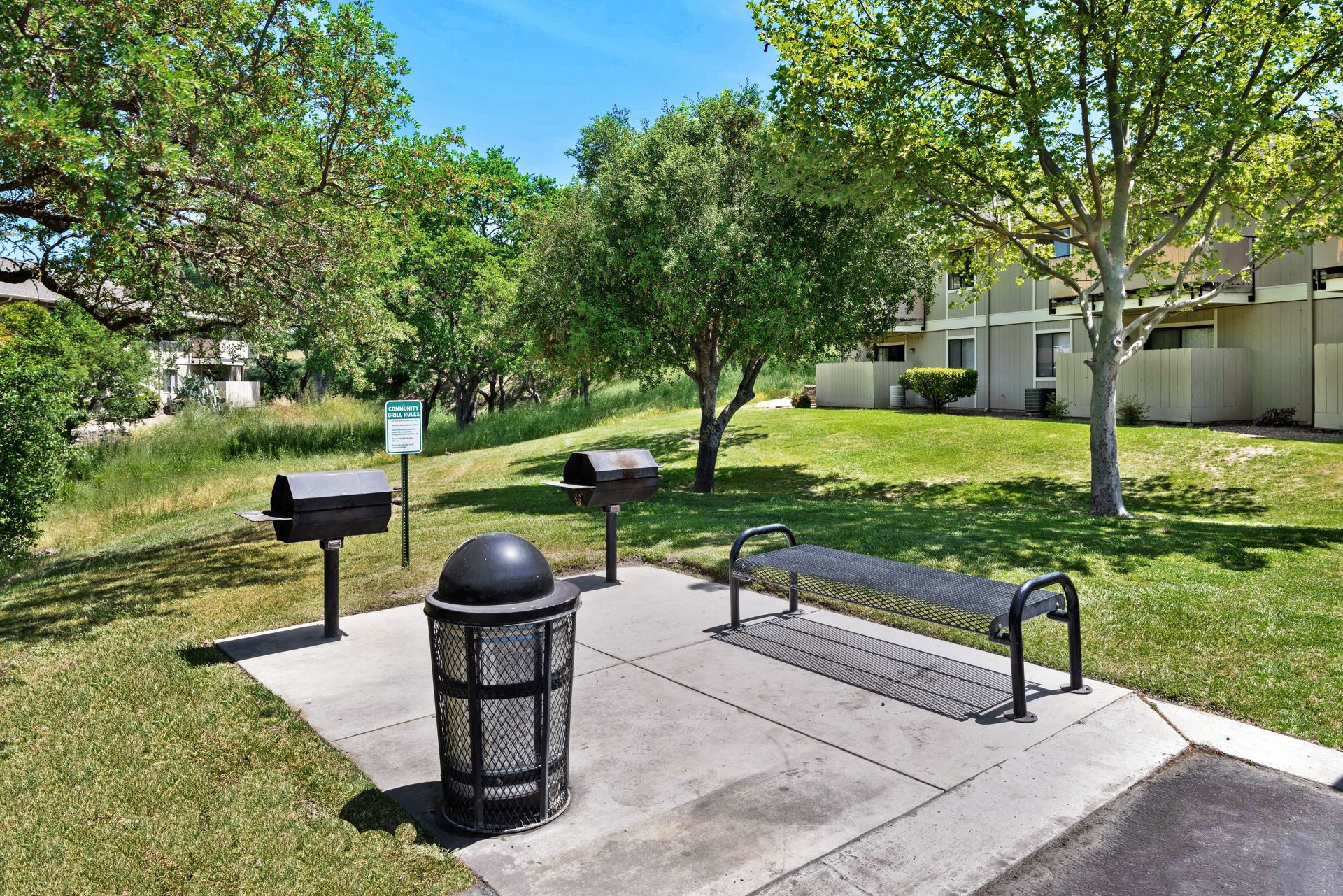 Park area with grills, a bench, and trash can in front of an apartment complex.