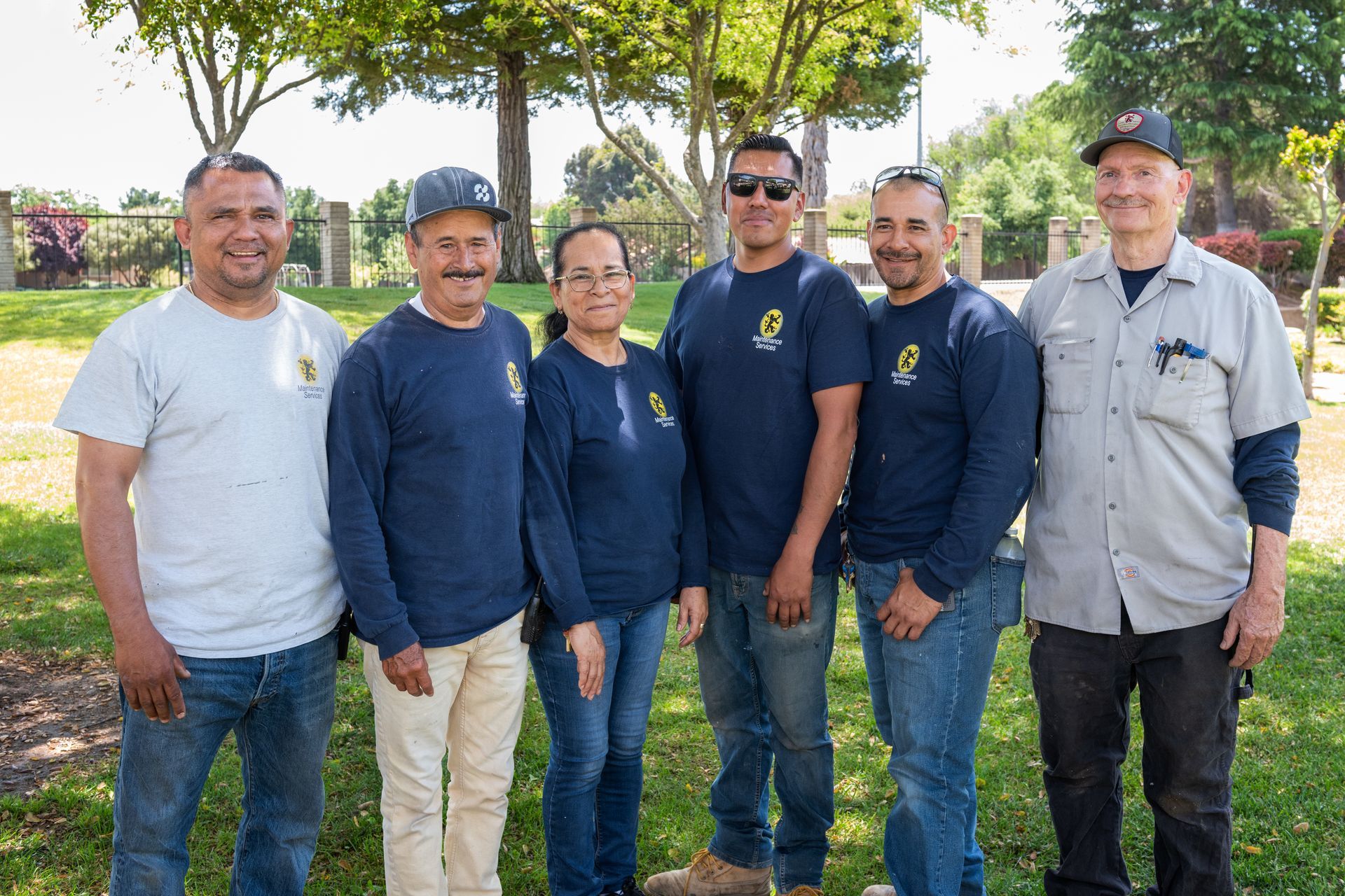 Group of six people standing outdoors in front of trees and grass. They are wearing work shirts and jeans.