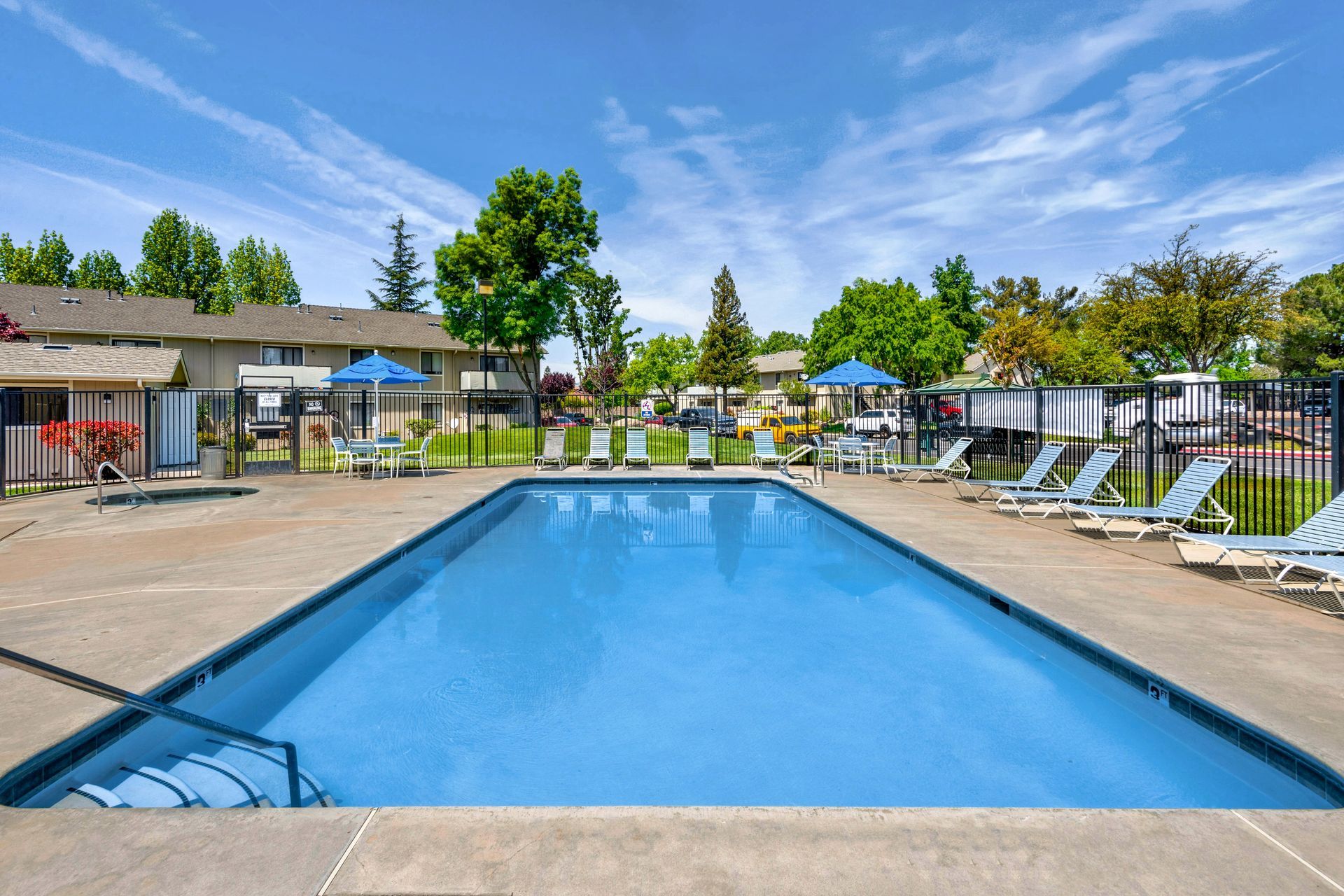 Pool with blue water, lounge chairs, and umbrellas on a sunny day in front of apartments.