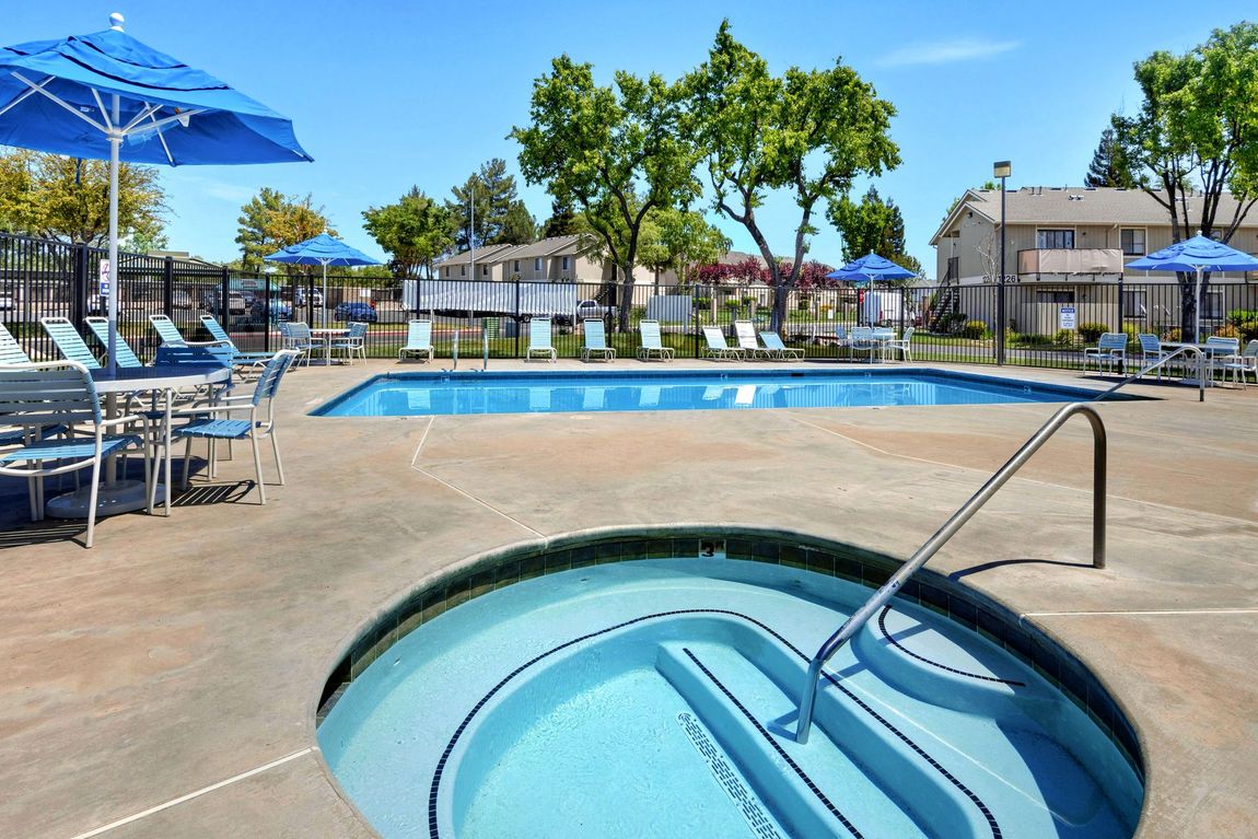 Pool area with a hot tub in the foreground, blue umbrellas, and lounge chairs under a sunny sky.