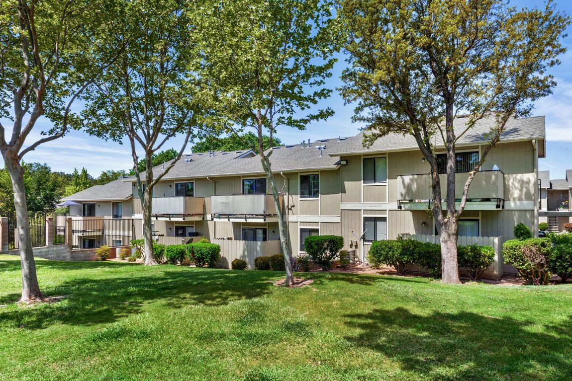 Two-story beige apartment building with balconies, trees, and green lawn on a sunny day.
