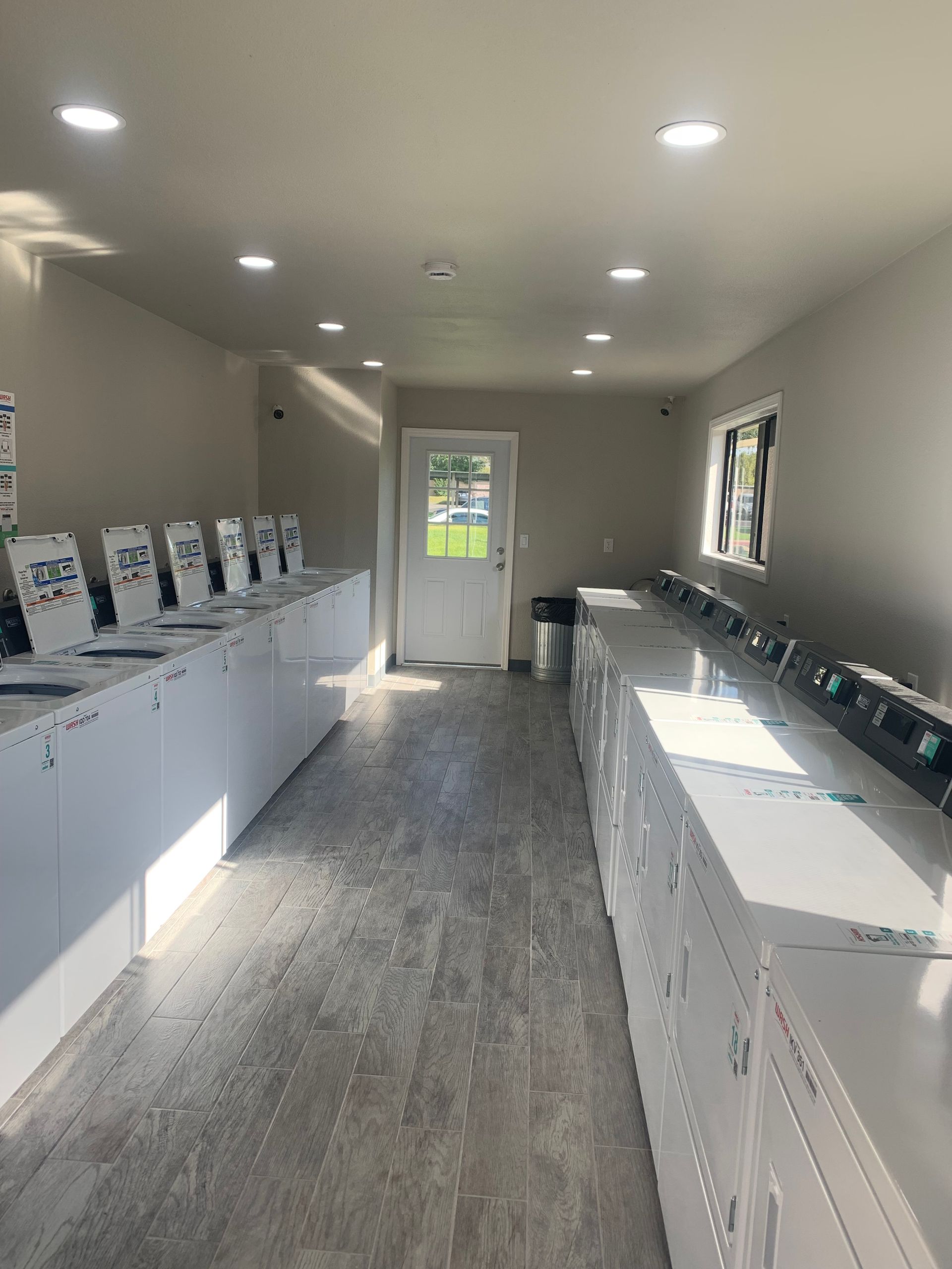 Interior of a laundromat with rows of white washing machines and dryers. White walls, gray floor, and a door at the end.