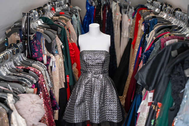 Woman in plaid coat, orange scarf, and sunglasses stands outside, holding her collar.