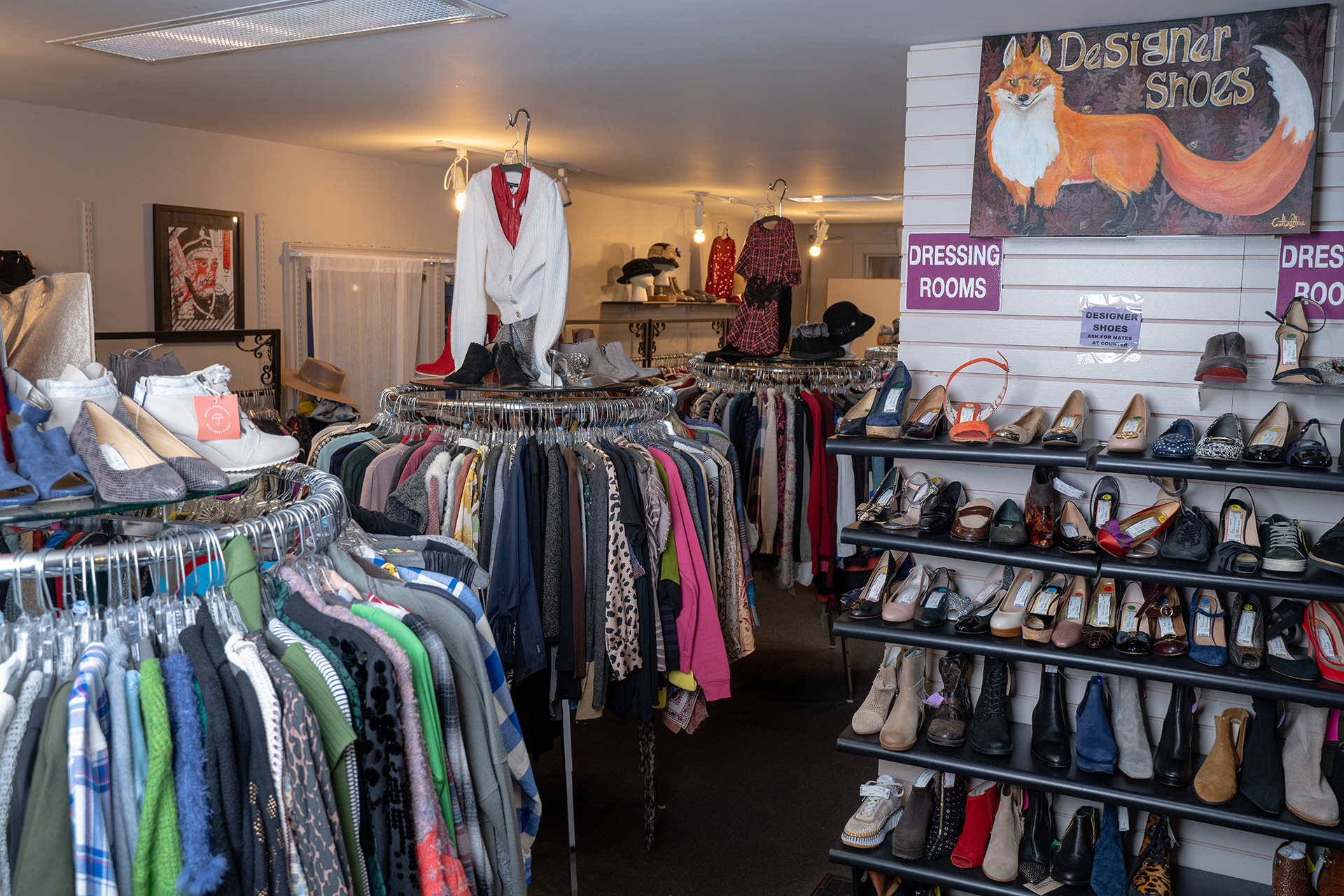 Two smiling women holding shoes in a store; one wearing a tan blazer, the other a turtleneck,