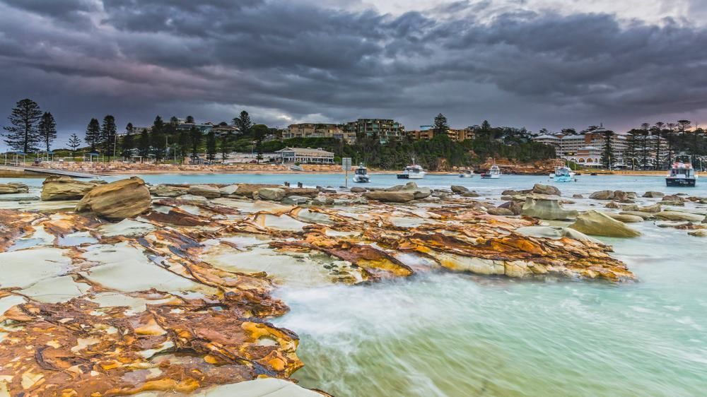 A Beach With Rocks and Boats in the Water on a Cloudy Day — Tuggerah Lakes Glass In Gosford, NSW
